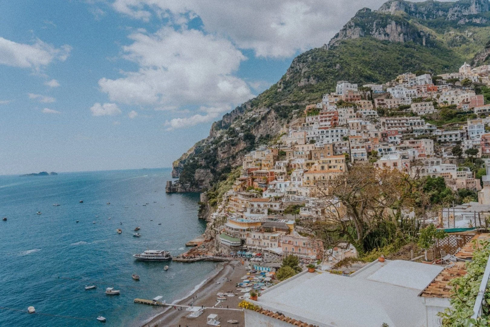 Cliffside view of Positano, Italy with colorful buildings cascading down the hillside to the beach and blue water, under partly cloudy sky.