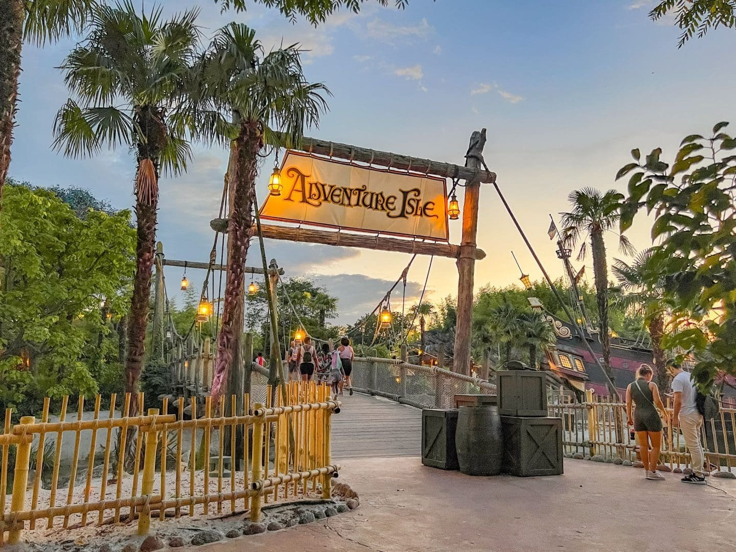 People walking on a wooden bridge at Adventure Isle during sunset, surrounded by lush green trees and pirate ships themed attractions.