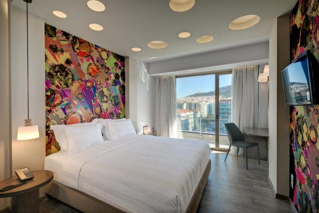 Hotel room with a large bed, colorful abstract artwork behind the bed, a sitting area with a view of city buildings and hills through large windows, and modern decor.