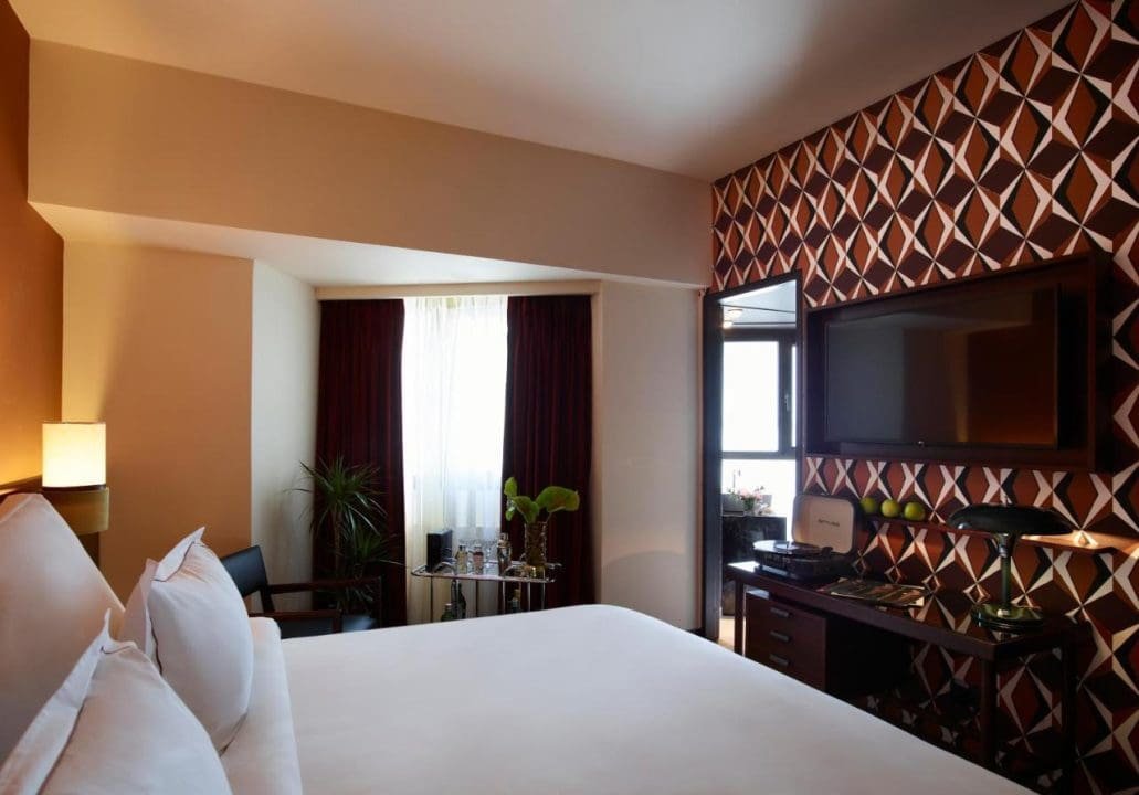 Hotel room with a large bed, a wall-mounted TV, a patterned accent wall, a window with curtains, a glass table with water bottles, plants, and a chair.