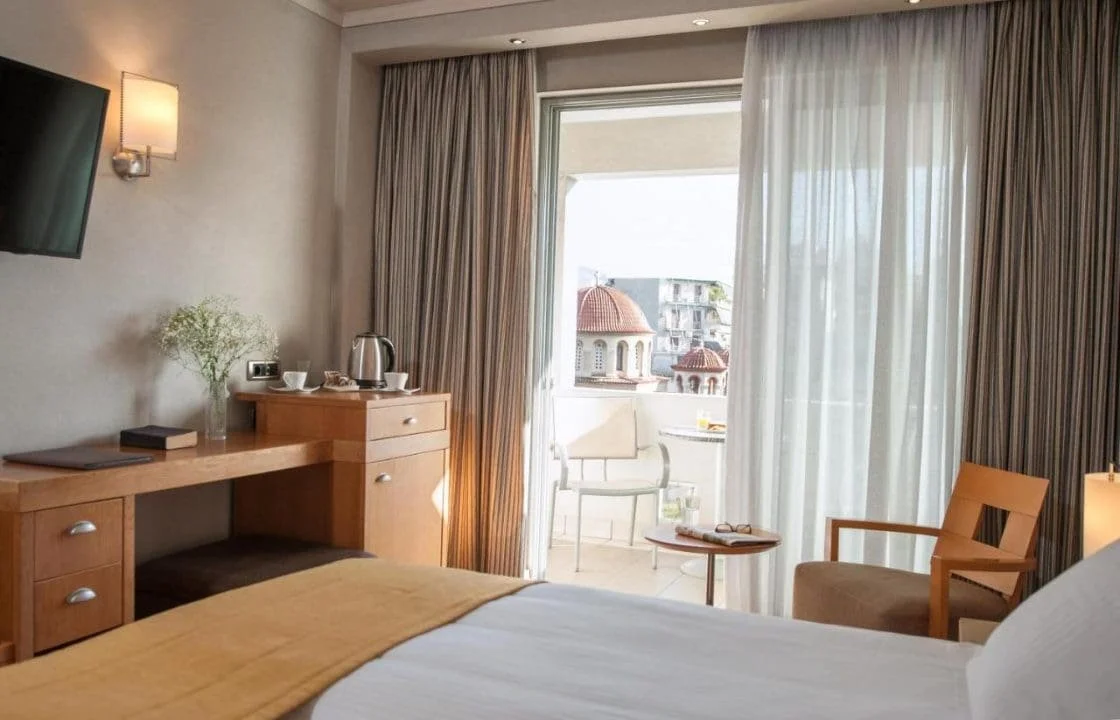 Hotel room with bed, wooden desk, flat-screen TV, balcony with outdoor seating, sheer curtains, and city view.