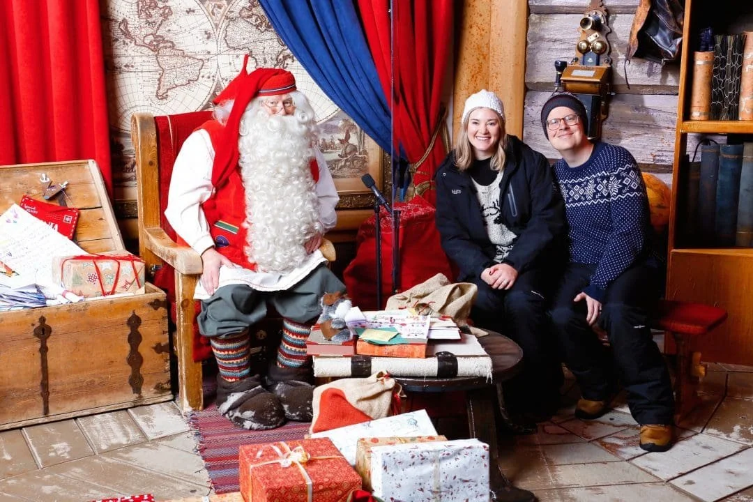Santa Claus sitting on a wooden chair surrounded by wrapped Christmas presents, with a woman and a man sitting beside him, all smiling. The room has red and blue curtains, a world map on the wall, and Christmas decorations.
