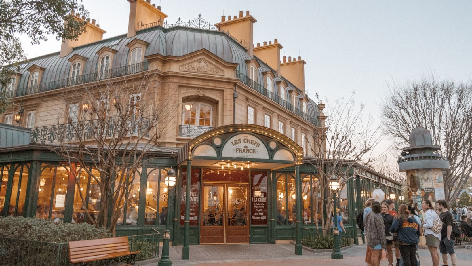 French restaurant called Les Chefs de France with people standing outside on a sidewalk, bare trees, and a multi-story building with a mansard roof in the background.