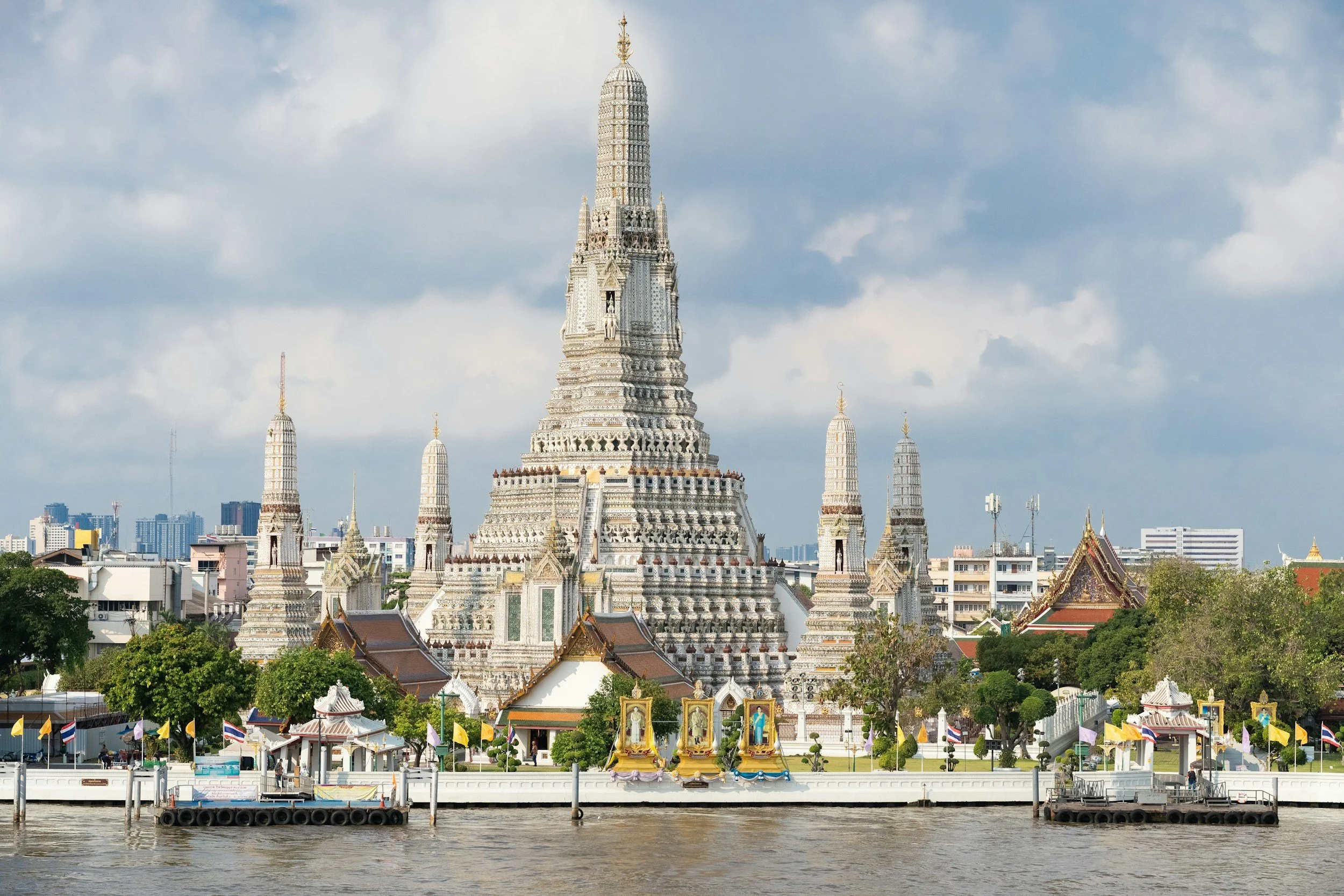 Wat Arun temple with a river in the foreground and skyscrapers in the background under a partly cloudy sky.