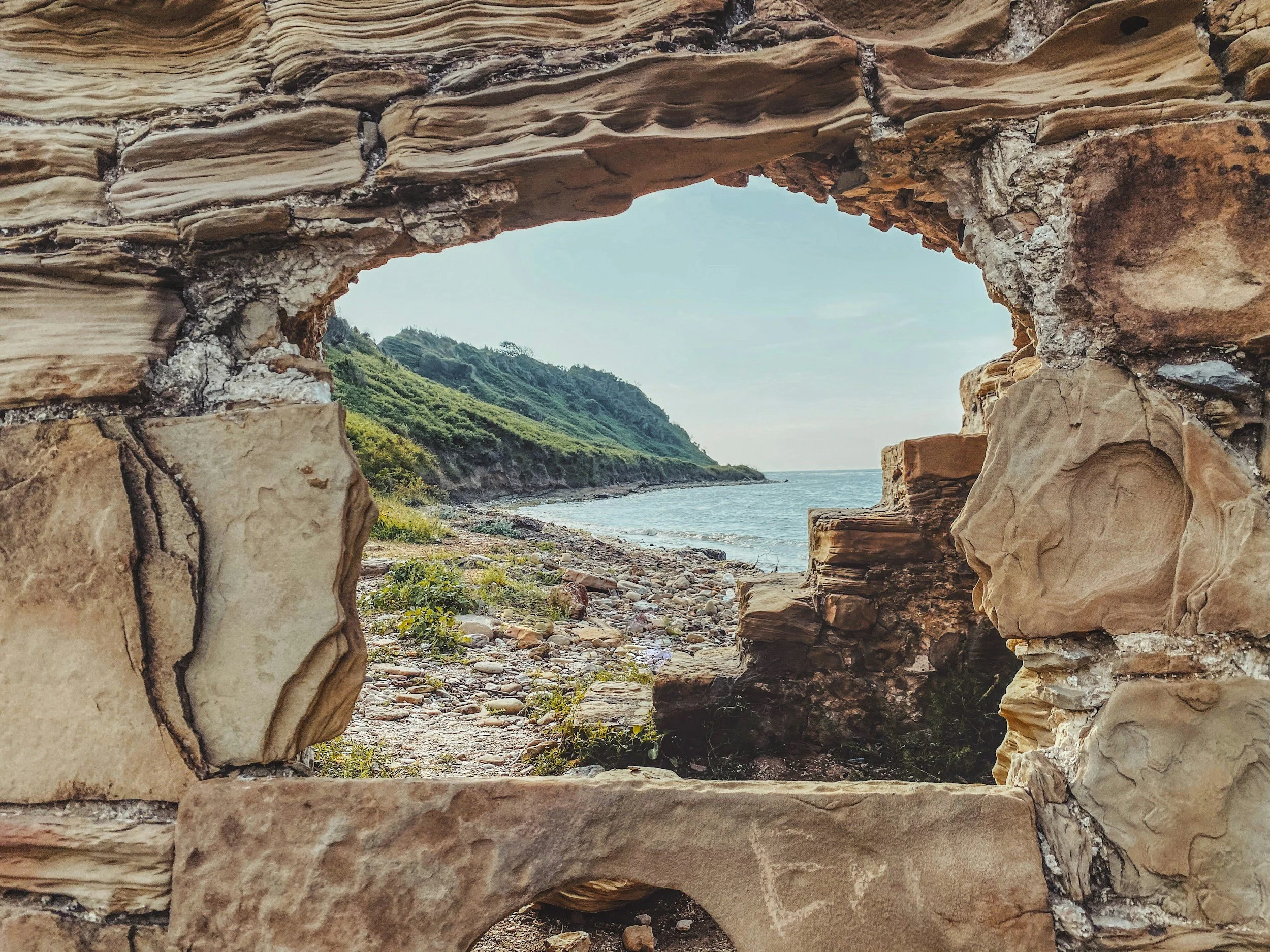 View of a rocky beach framed through a stone window, with a hill covered in greenery, a shoreline, and the ocean in the distance.