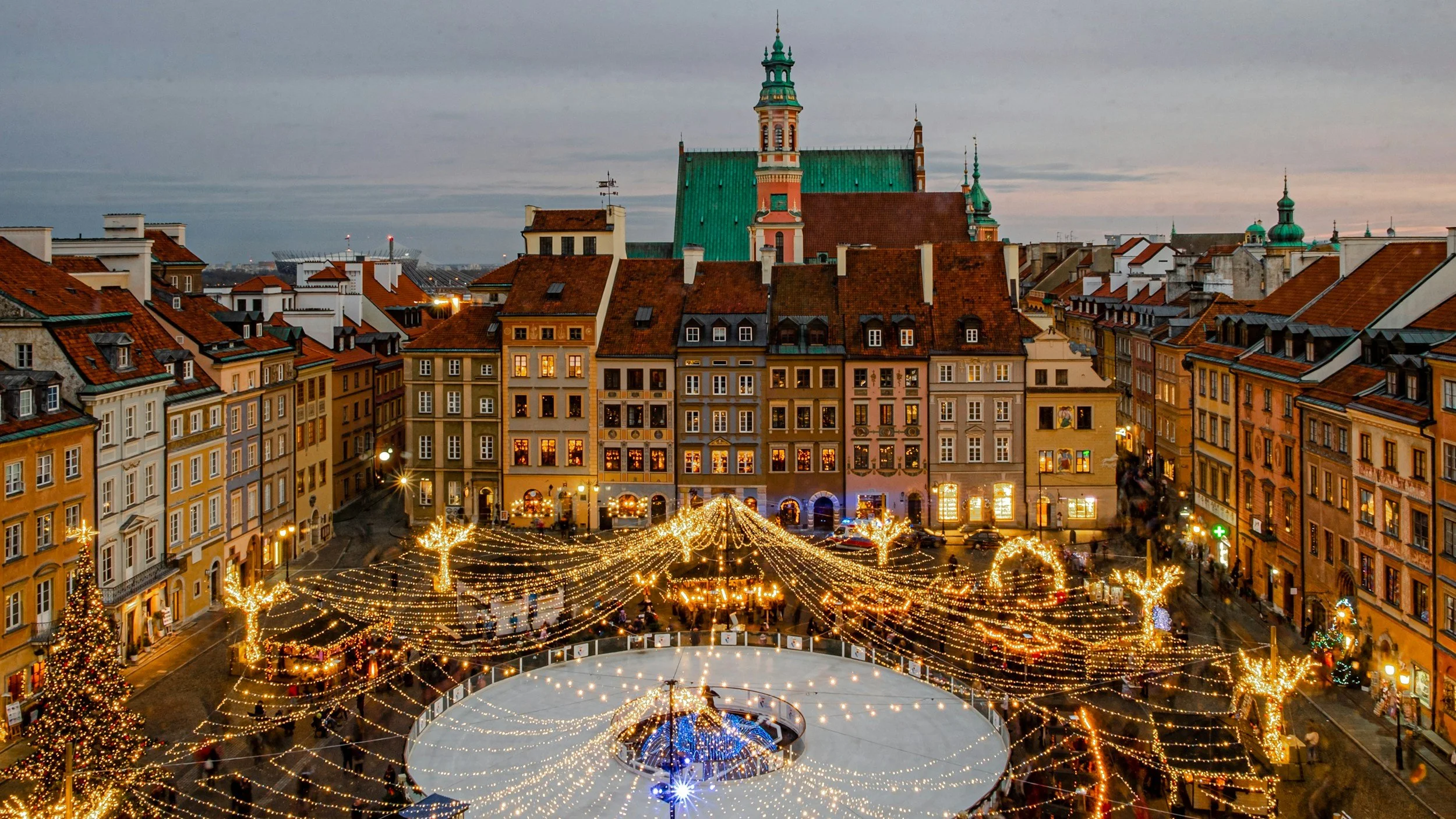 A European city square decorated with Christmas lights, illuminated trees, and a Christmas tree in the foreground, with historic buildings and church spires in the background during dusk.