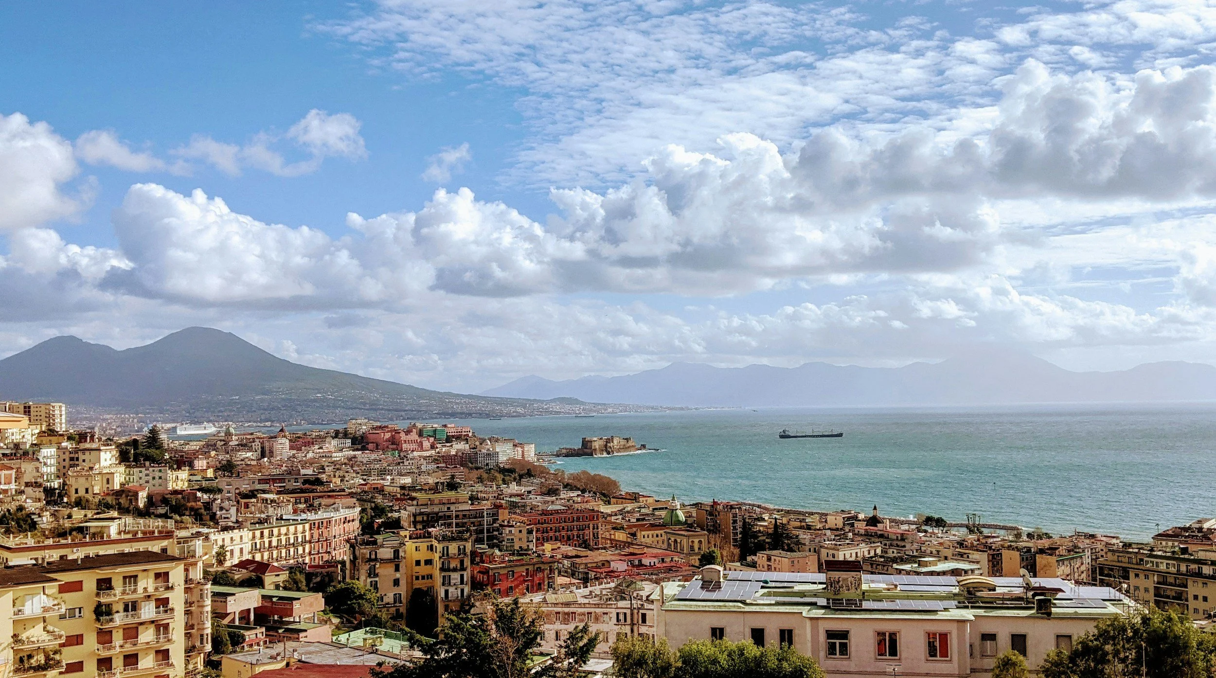 A cityscape with colorful buildings along the coast, mountains in the background, and a partly cloudy sky.