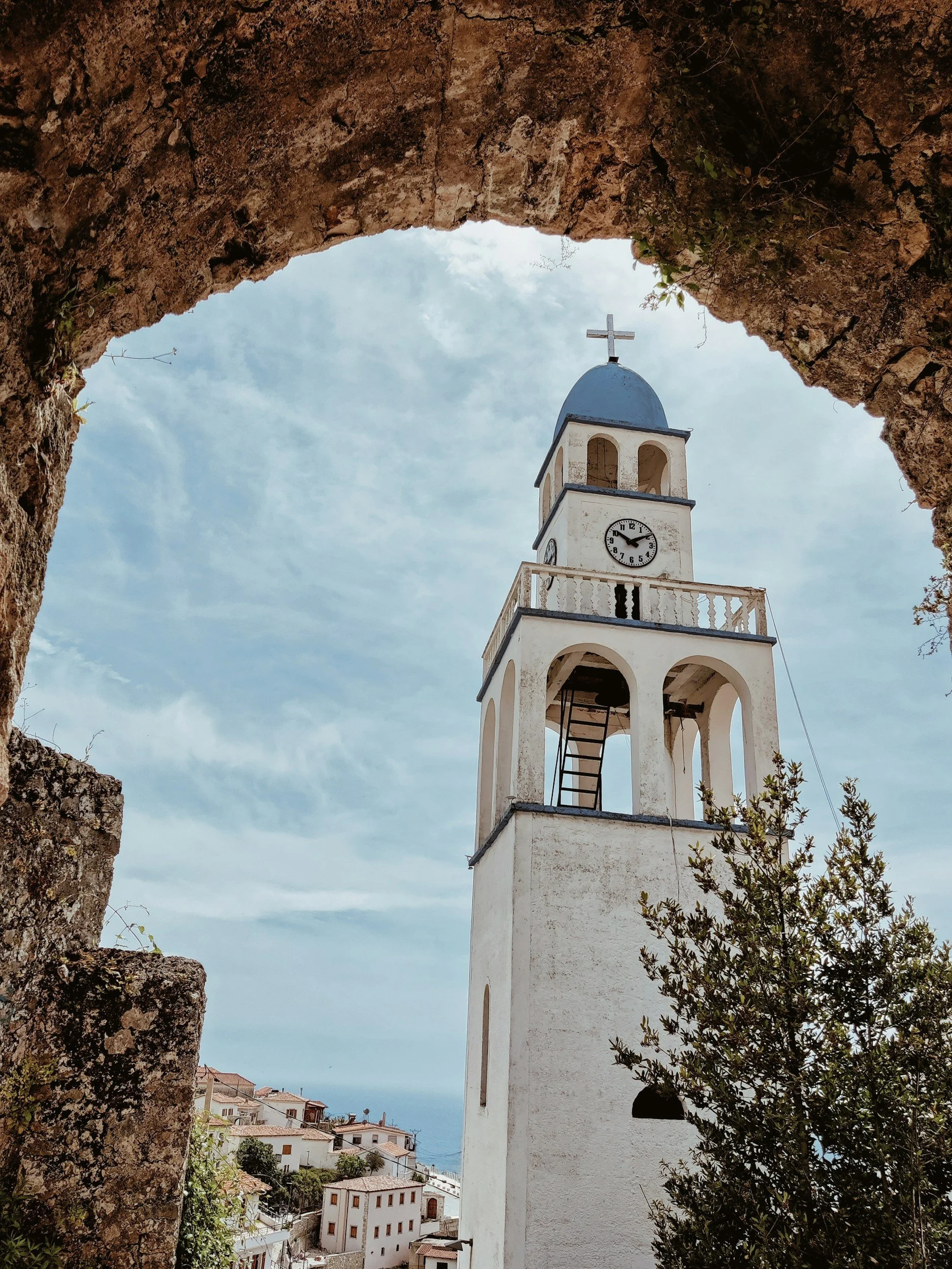 View of a white church tower with a clock and cross, framed by a stone archway, with buildings and the sea in the background.