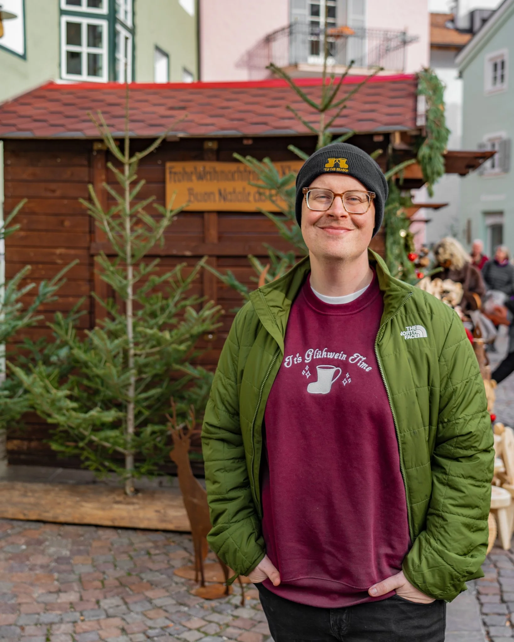 A man with glasses, a black beanie, a green jacket, and a maroon T-shirt with German text, standing outdoors in front of a small wooden Christmas market stall decorated with greenery and a sign, with a colorful European-style street scene in the background.