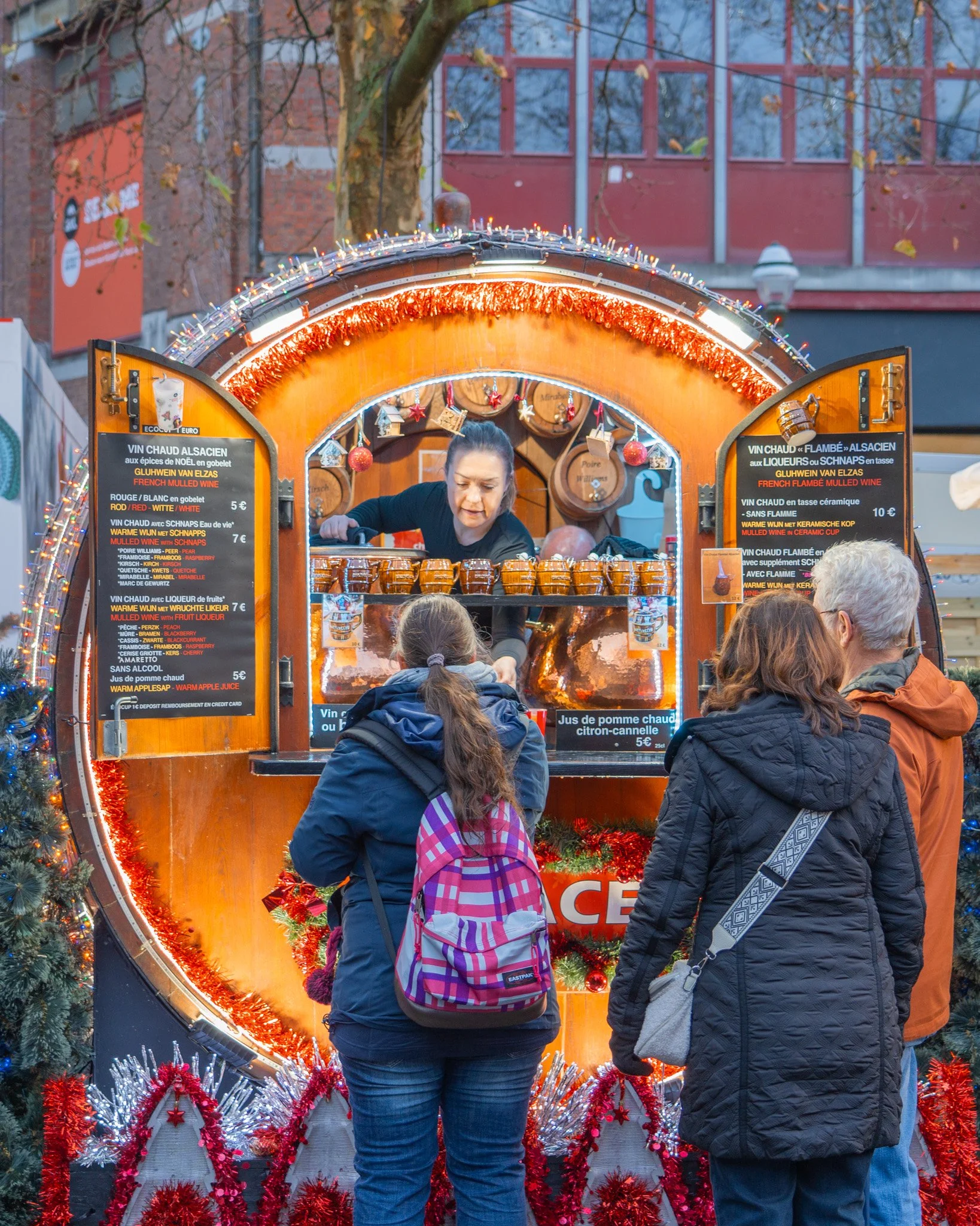 People standing in front of a festive, decorated Christmas market stall selling mulled wine, with Christmas ornaments, lights, and tinsel.