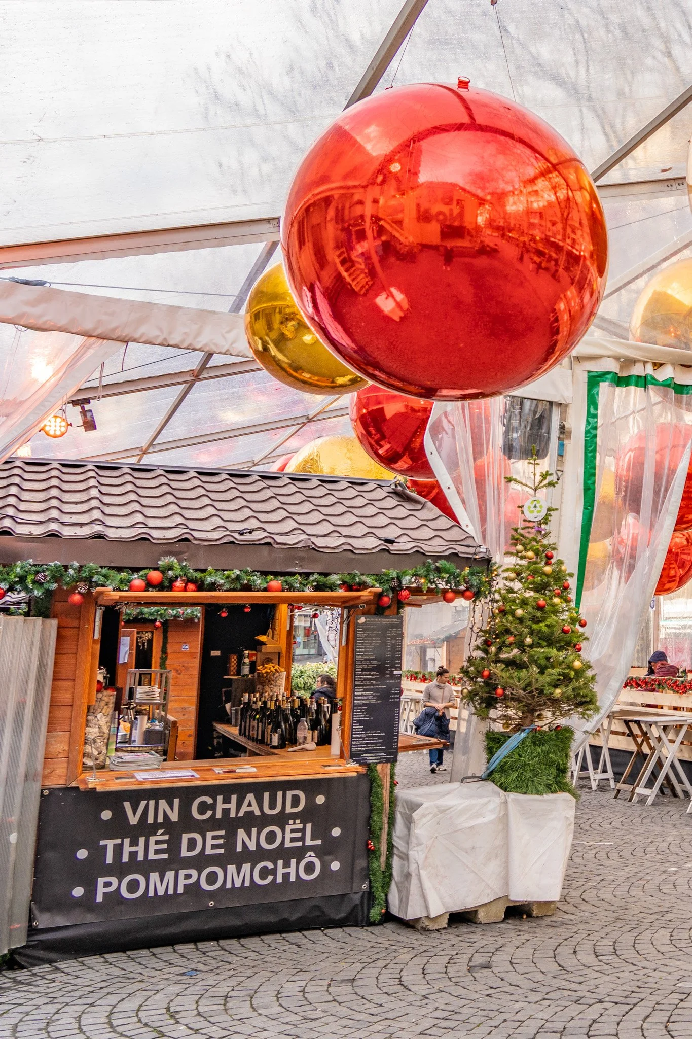 Christmas market stall decorated with garland and ornaments selling hot drinks and wine, with Christmas trees nearby. Large red and gold Christmas ornaments hanging from the ceiling.