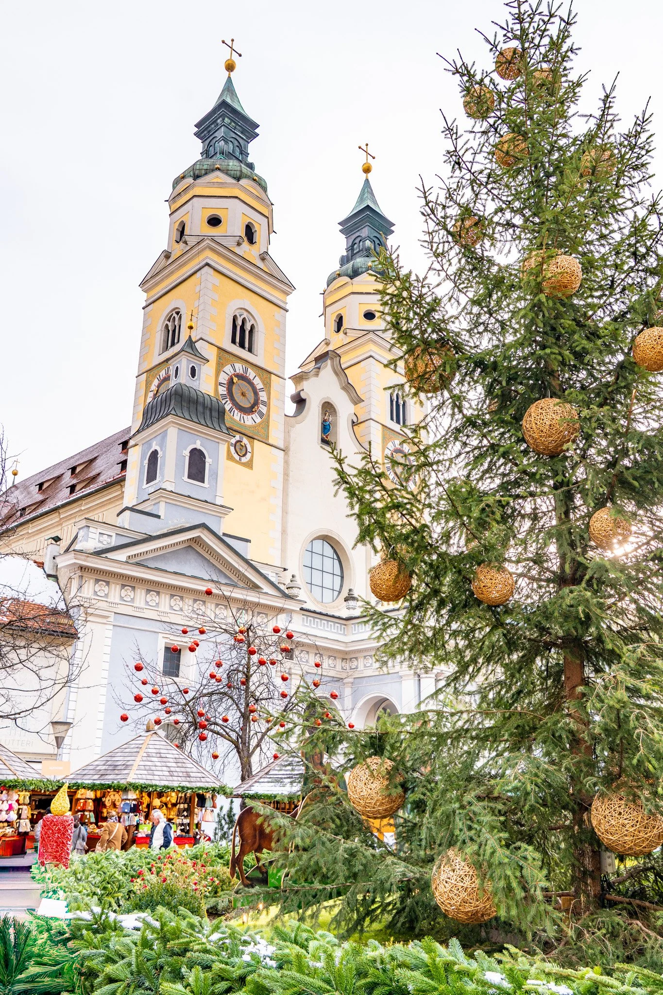 Decorated Christmas tree with woven golden ornaments in front of a historic church with twin towers and a Christmas market with stalls and visitors.