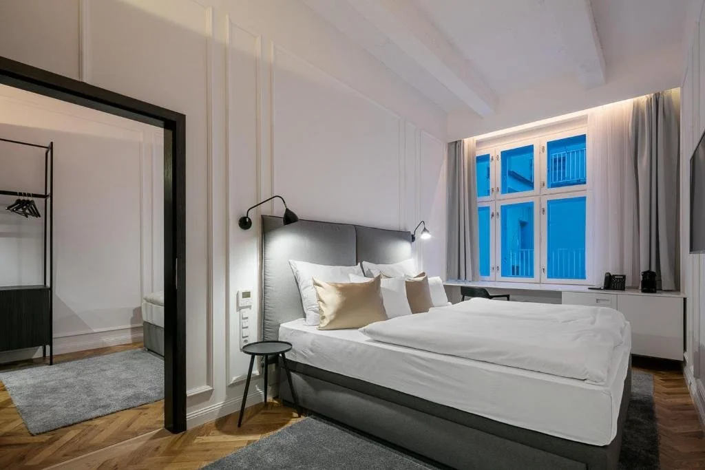 A modern hotel room with a large bed, bedside lamps, a window with curtains, and a small workspace. The room has white walls and wooden flooring.