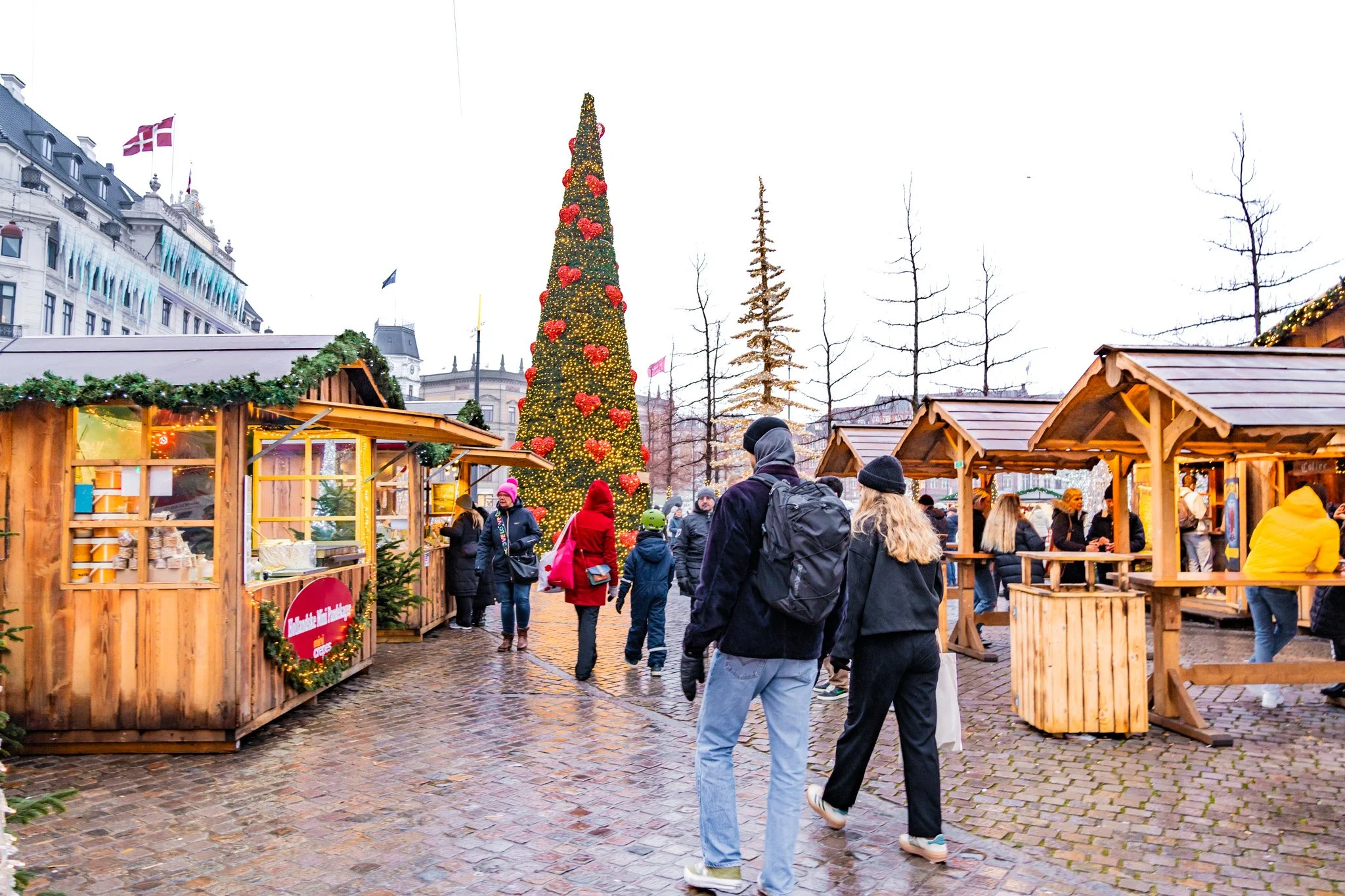 A small Christmas-themed food kiosk with festive decorations, surrounded by snow-dusted trees and plants, with a sign reading 'Nissens Rugbyrossandwich' and another sign 'BJÆLDEVANG', in an outdoor holiday market setting.