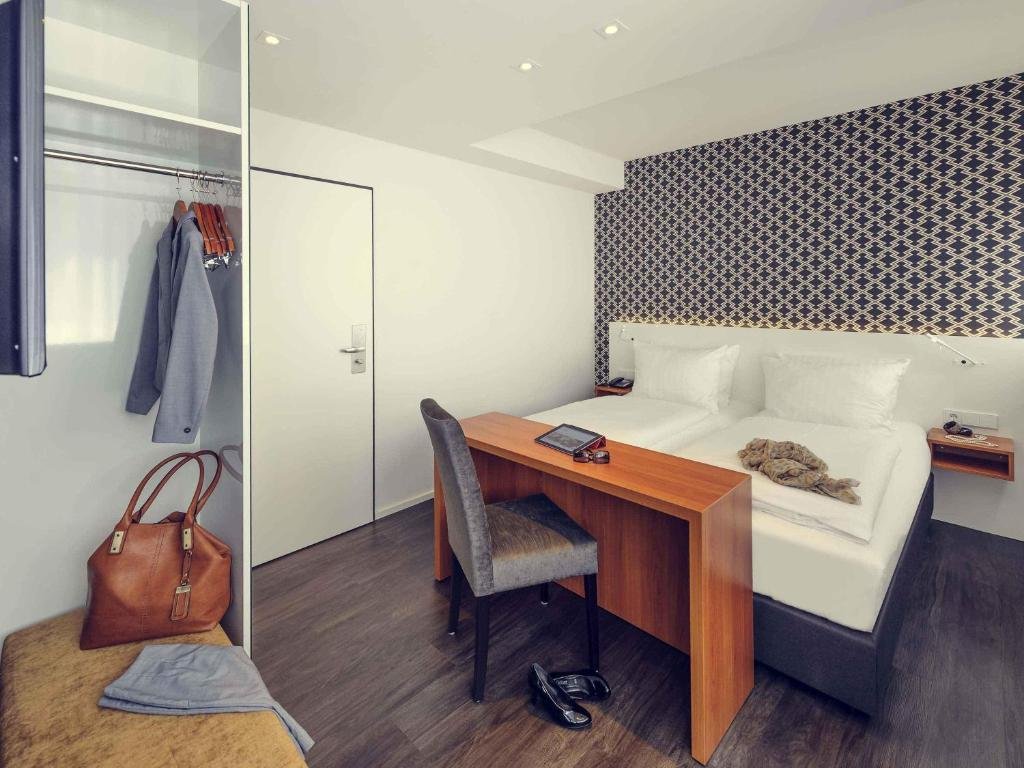 Hotel room with a white bed, a wooden desk, and a gray upholstered chair. A decorative patterned accent wall behind the bed, with a bedside table and phone. Closet with clothes and a large brown handbag, and a small bench with a towel on top.
