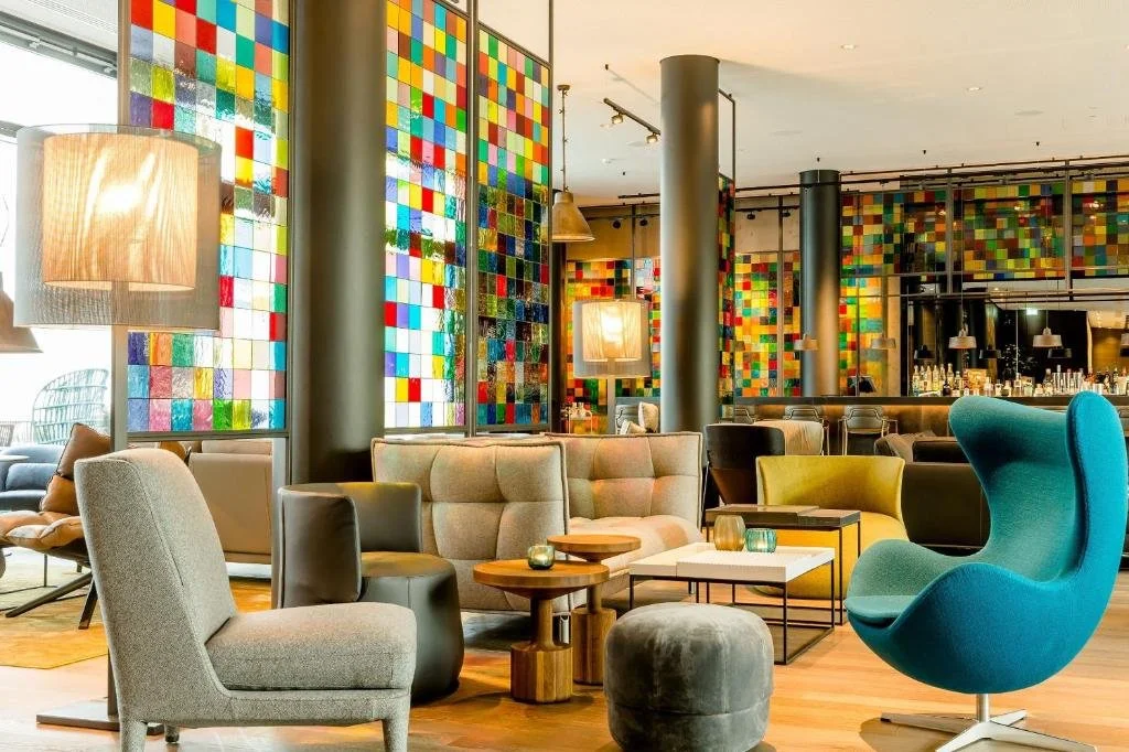 Interior of a modern lounge with colorful stained glass windows, various stylish armchairs, a bar in the background, and decorative lighting.