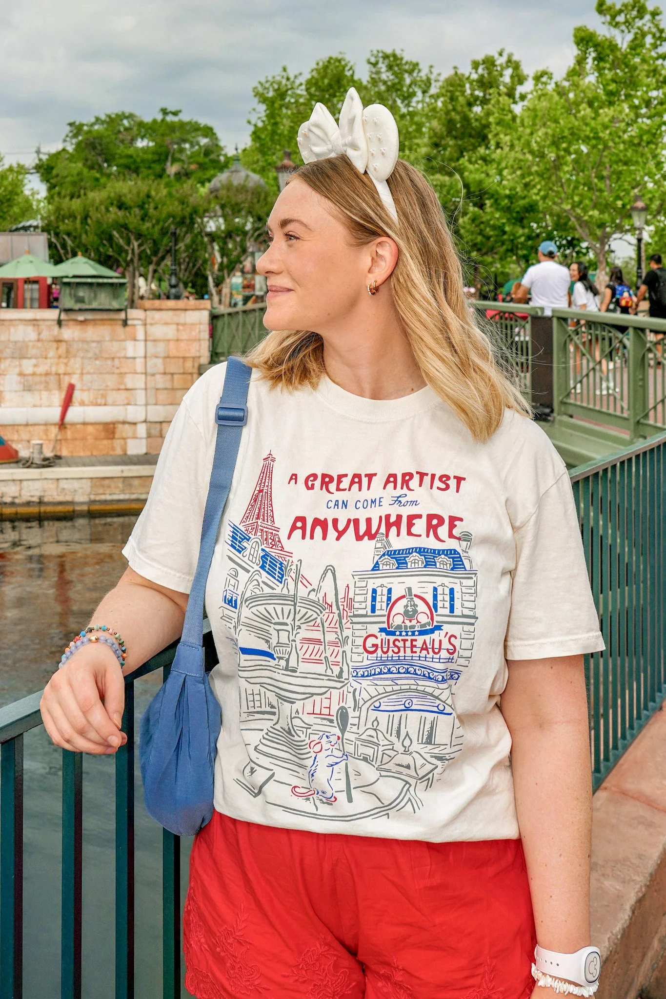 Close-up of a woman with blonde hair wearing a white T-shirt with red and blue print, red shorts, a blue bag, and accessories including a white headband with a bow, colorful bracelets, and a white watch, standing outdoors near a green railing over water with green trees and people in background.