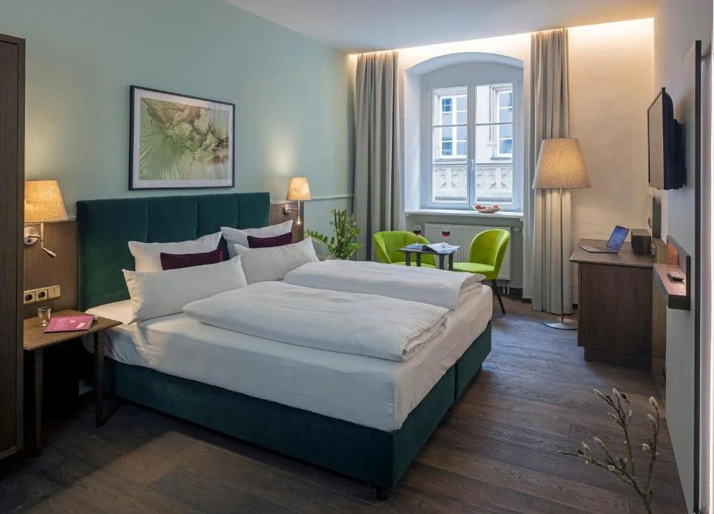 A hotel room with a large bed, green headboard, white bedding, and purple pillows. There are two lamps on each side of the bed, a window with curtains, a small table with two green chairs, a wooden cabinet with a TV, and framed artwork above the bed.