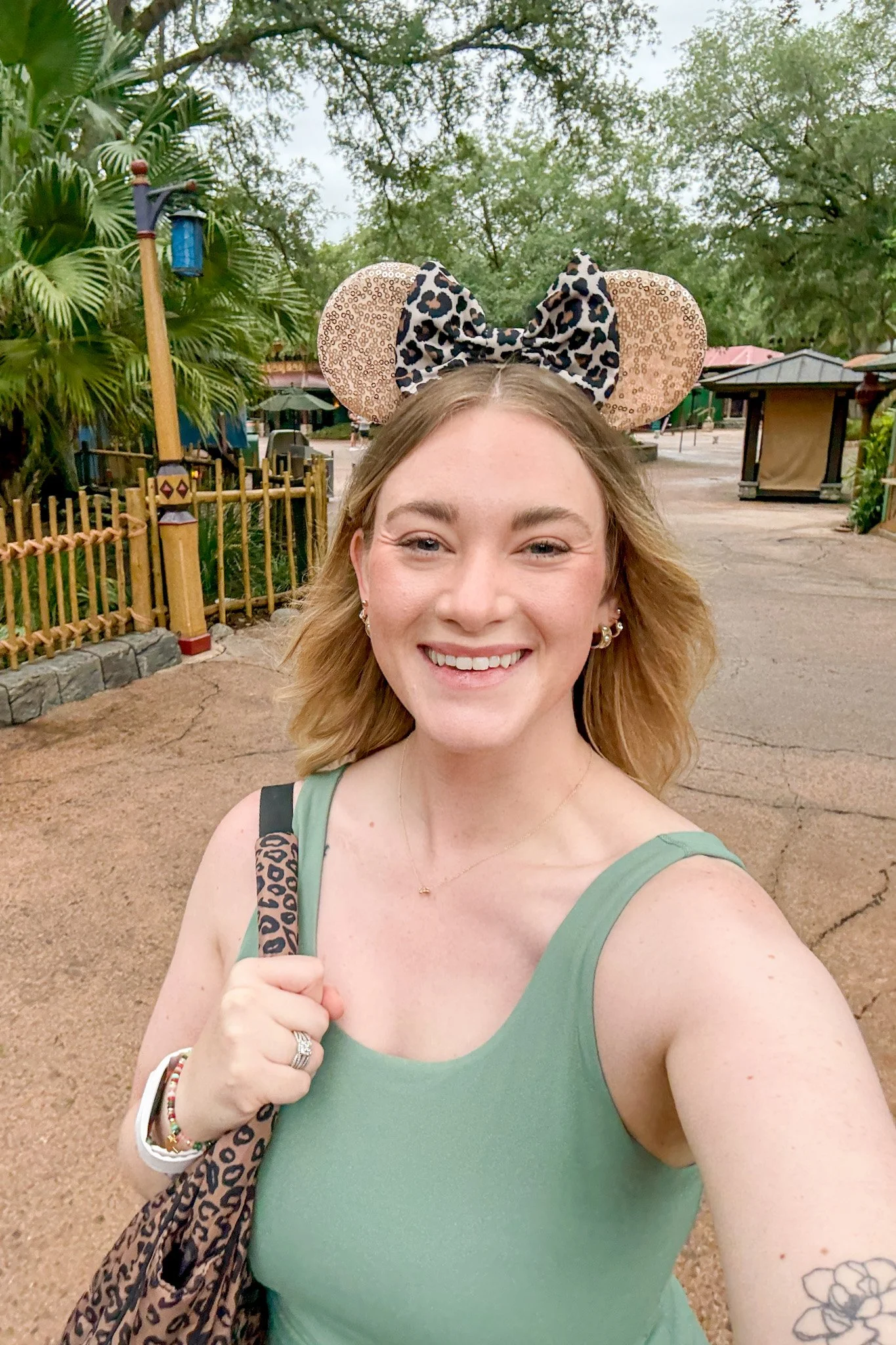 A smiling young woman with shoulder-length blonde hair wearing a green tank top, Mickey Mouse ears with a leopard print bow, and a leopard print backpack, taking a selfie outdoors in a themed park with trees, a wooden fence, and park booths in the background.