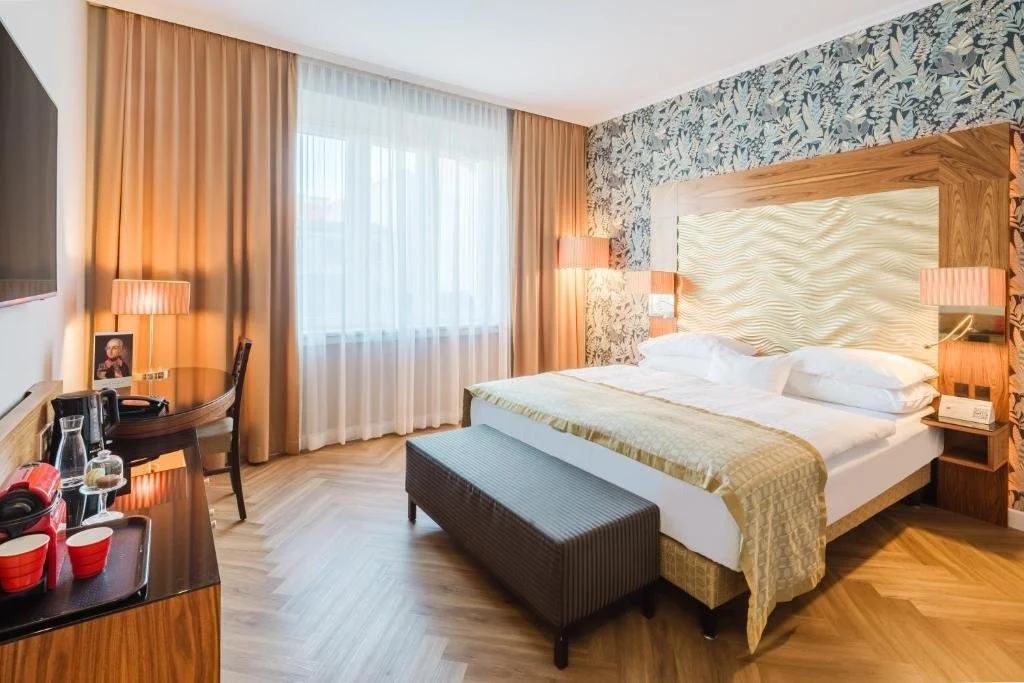Modern hotel bedroom with large bed, wooden headboard, floral wallpaper, and floor-length curtains, with bedside lamps, a bench at the foot of the bed, and a wooden desk with refreshments.