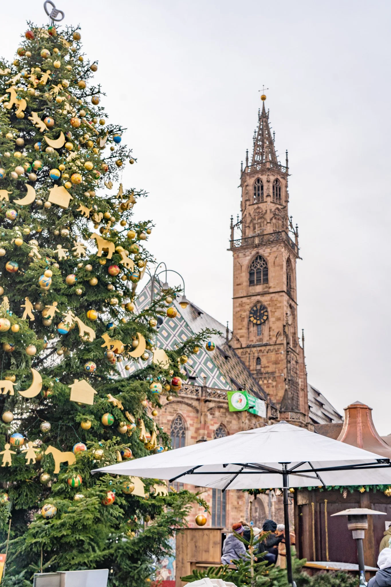 A decorated Christmas tree with gold, green, and red ornaments, gold wooden cutouts, and a star topper, set outdoors near a stone church with a tall spire. There are umbrellas and people gathered around, some wearing winter clothing.