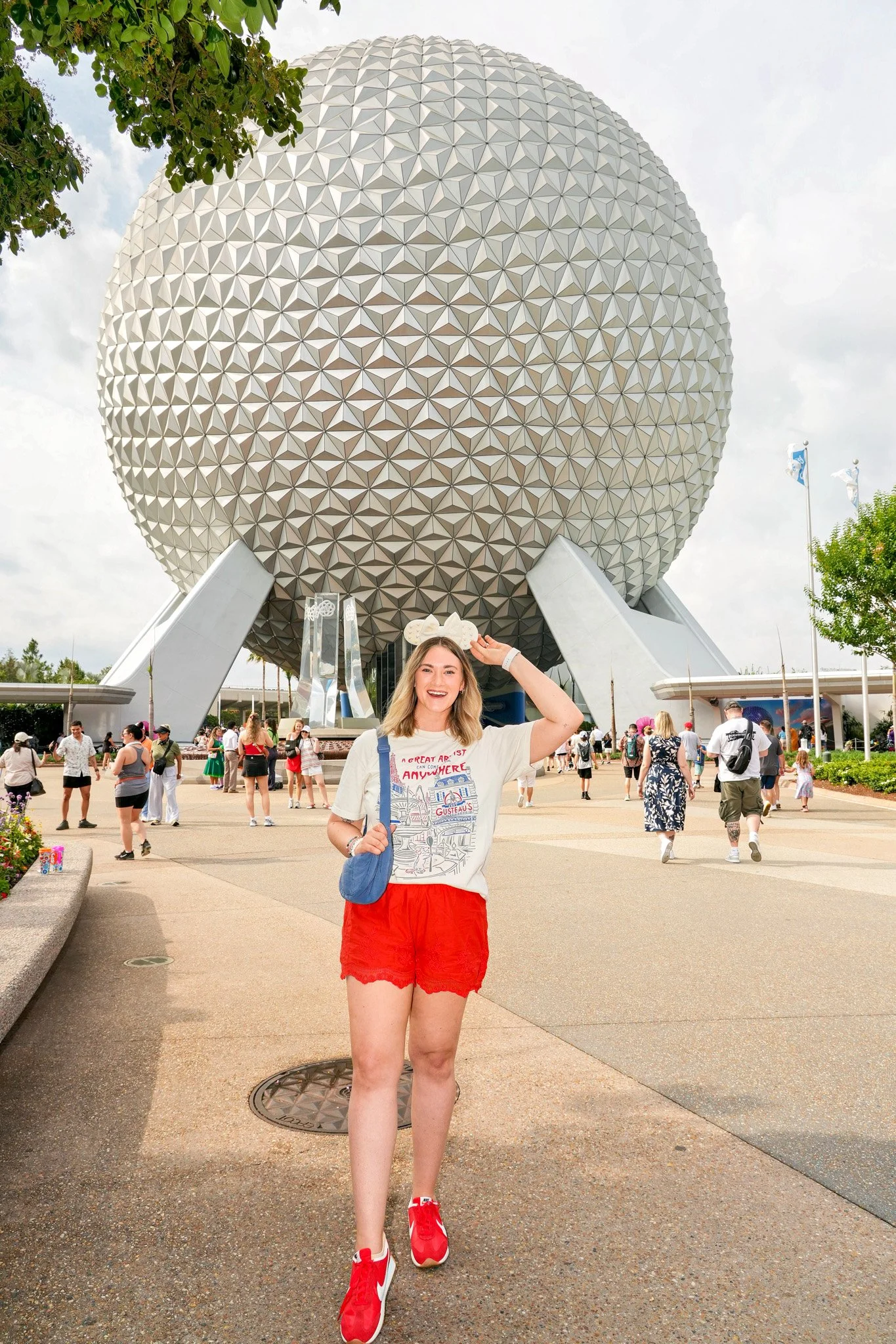 A young woman with long blonde hair, smiling, wearing a white T-shirt, red shorts, and red sneakers, standing in front of Spaceship Earth at EPCOT theme park at Walt Disney World.