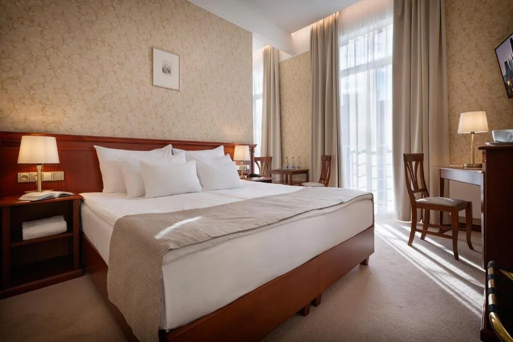 A hotel room with a large bed, two bedside tables with lamps, curtains over a balcony door, a table with two glasses, and a TV mounted on the wall.