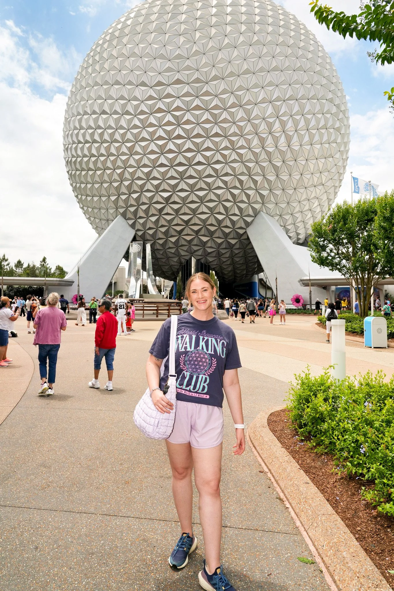 A smiling woman in a gray t-shirt and pink shorts walking in front of the Spaceship Earth structure at Epcot, Disney World, with tourists around and blue sky with some clouds overhead.