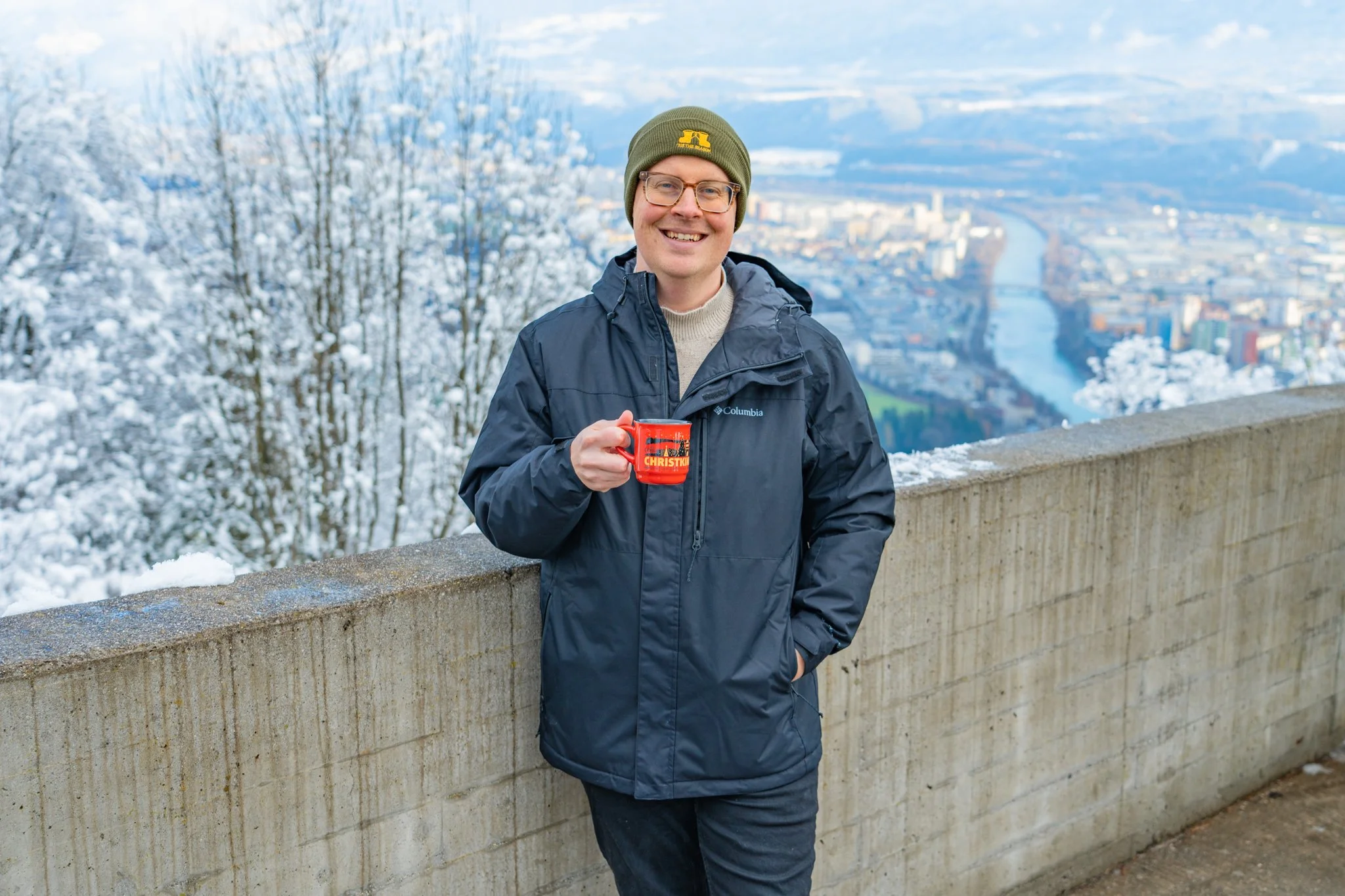 Man smiling and holding a red mug labeled 'Christmas' while standing outdoors on a snowy day with a city view in the background.