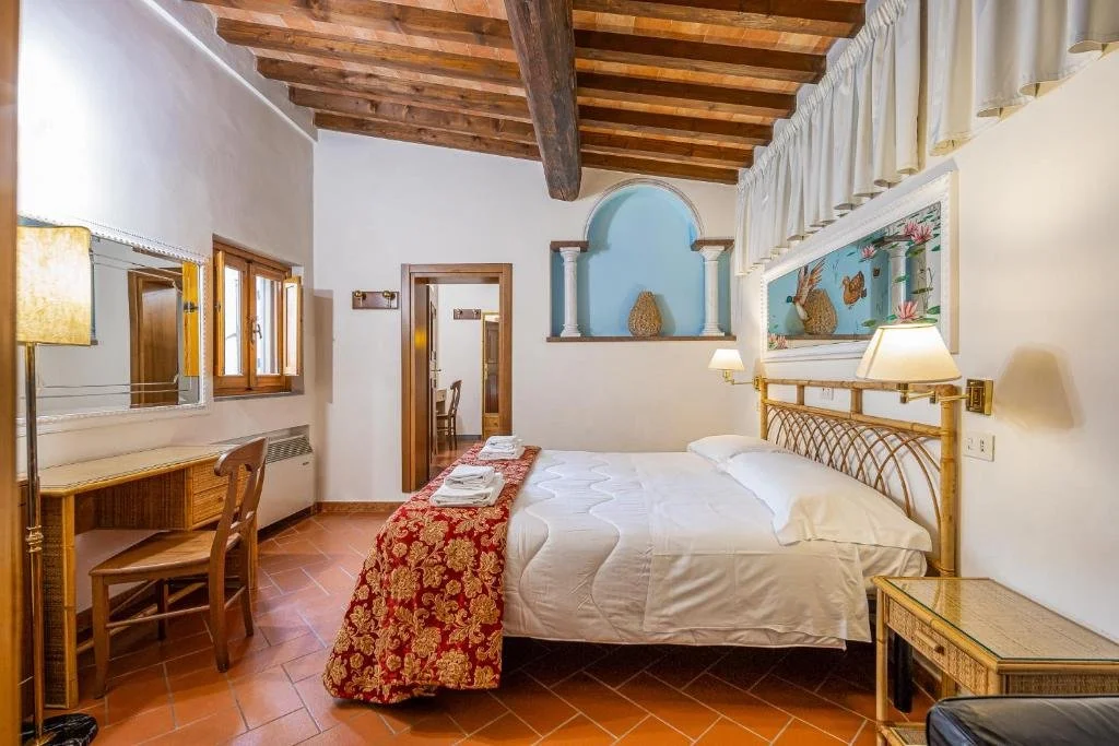 A cozy hotel room with a brass bed, white bedding, and a red and gold patterned throw. There are wall lamps on each side of the bed, a large framed painting above, wood ceiling beams, a small desk with a chair, and a window with wooden shutters. An open door leads to another room.