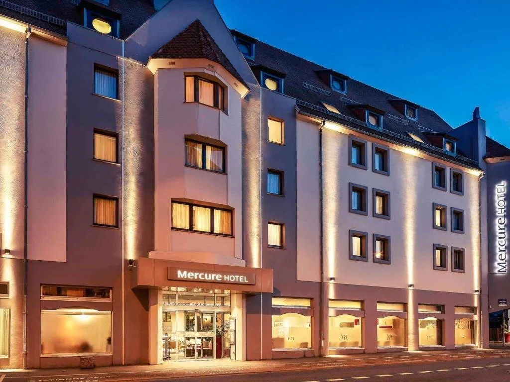 Exterior view of the Mercure Hotel at dusk, showing its illuminated facade with multiple stories, a central entrance, and several windows with warm lighting.
