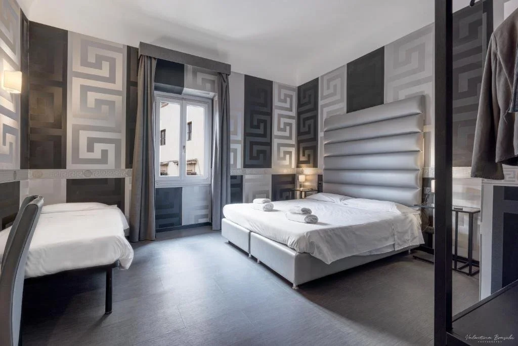 Modern hotel room with a large bed, decorative grey patterned wallpaper, a window with grey curtains, and a small bed or couch with white linens and towels.