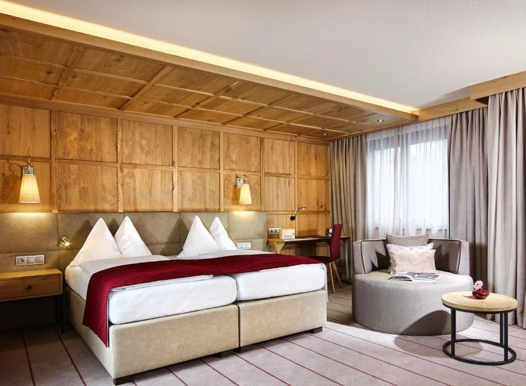 A hotel room with a double bed, beige headboard, wooden panel wall, two wall-mounted lamps, a small desk with a red chair, round gray armchair with pillows, round side table, and large windows with beige curtains.