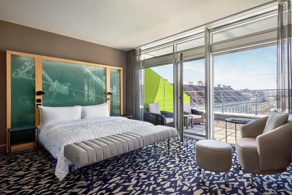Modern hotel room with a large bed, a bench, and seating area with two chairs and a small table. Floor-to-ceiling windows open to a balcony with outdoor seating and city views.
