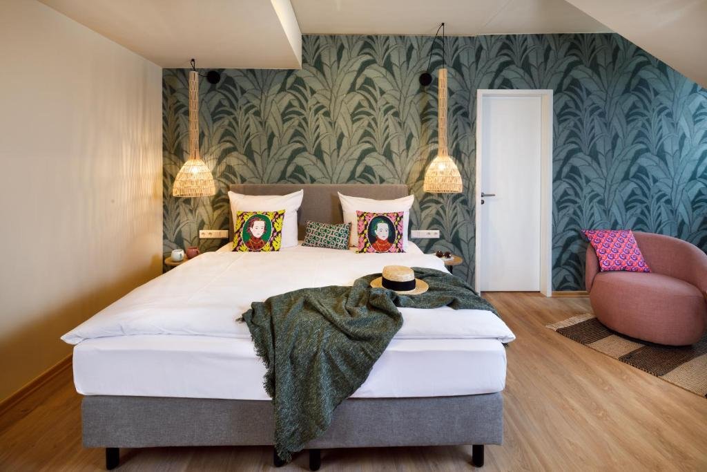 A bedroom with a large bed, decorative pillows, hanging pendant lights, a pink armchair, and wooden floor, featuring a patterned accent wall.
