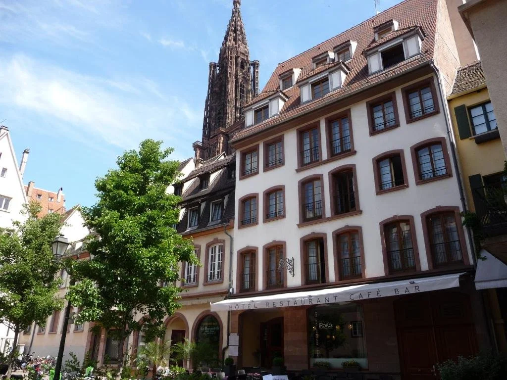 A European city street with a tall church spire in the background, and lined with traditional buildings. One building has a white facade with multiple windows and a sign indicating a hotel, restaurant, cafe, and bar. There are green trees and outdoor seating in the foreground.