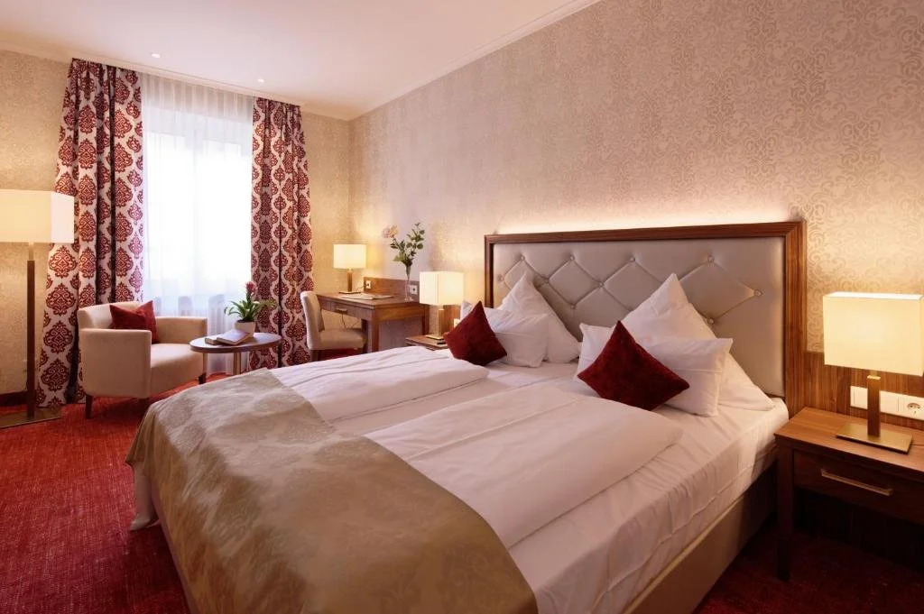 A hotel room with a large bed, patterned curtains, bedside lamps, a small armchair with a side table, a desk with a chair, and a flower vase, all in warm lighting.