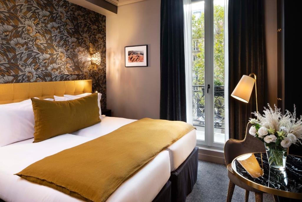 Hotel room with a bed featuring a mustard-colored headboard, blanket, and pillow, dark floral-patterned wallpaper, a small table with a black marble top, a vase of white flowers, a window with black curtains, a chair, and a standing floor lamp.