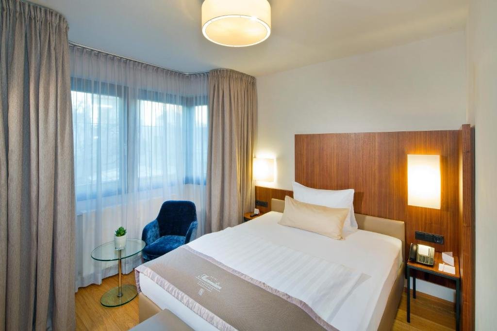 Hotel room with a large bed, wooden headboard, bedside lamps, a small glass table with a potted plant, a blue armchair, and large windows with sheer curtains.