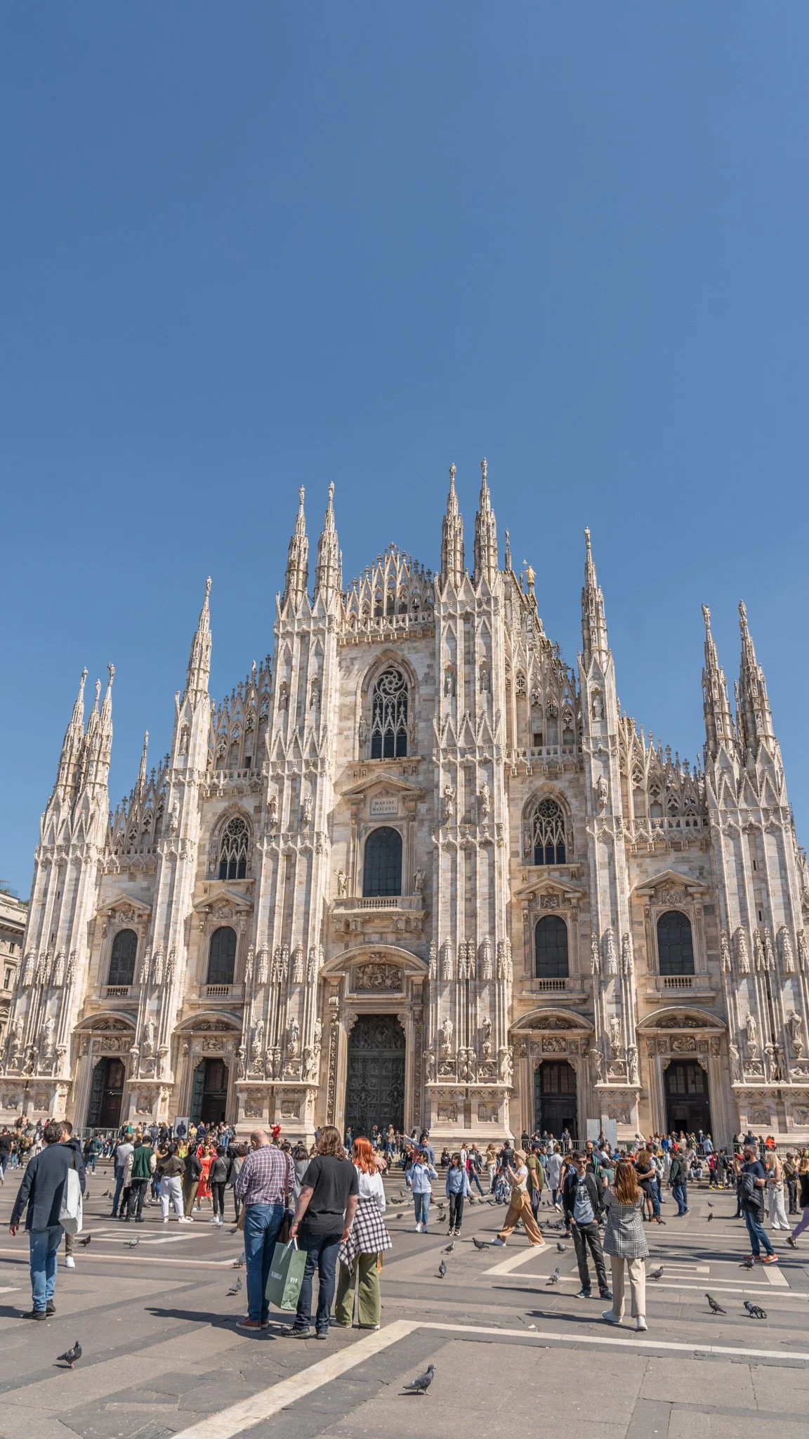 Front view of the Milan Cathedral (Duomo di Milano) in Italy, with people and pigeons in the square under a clear blue sky.