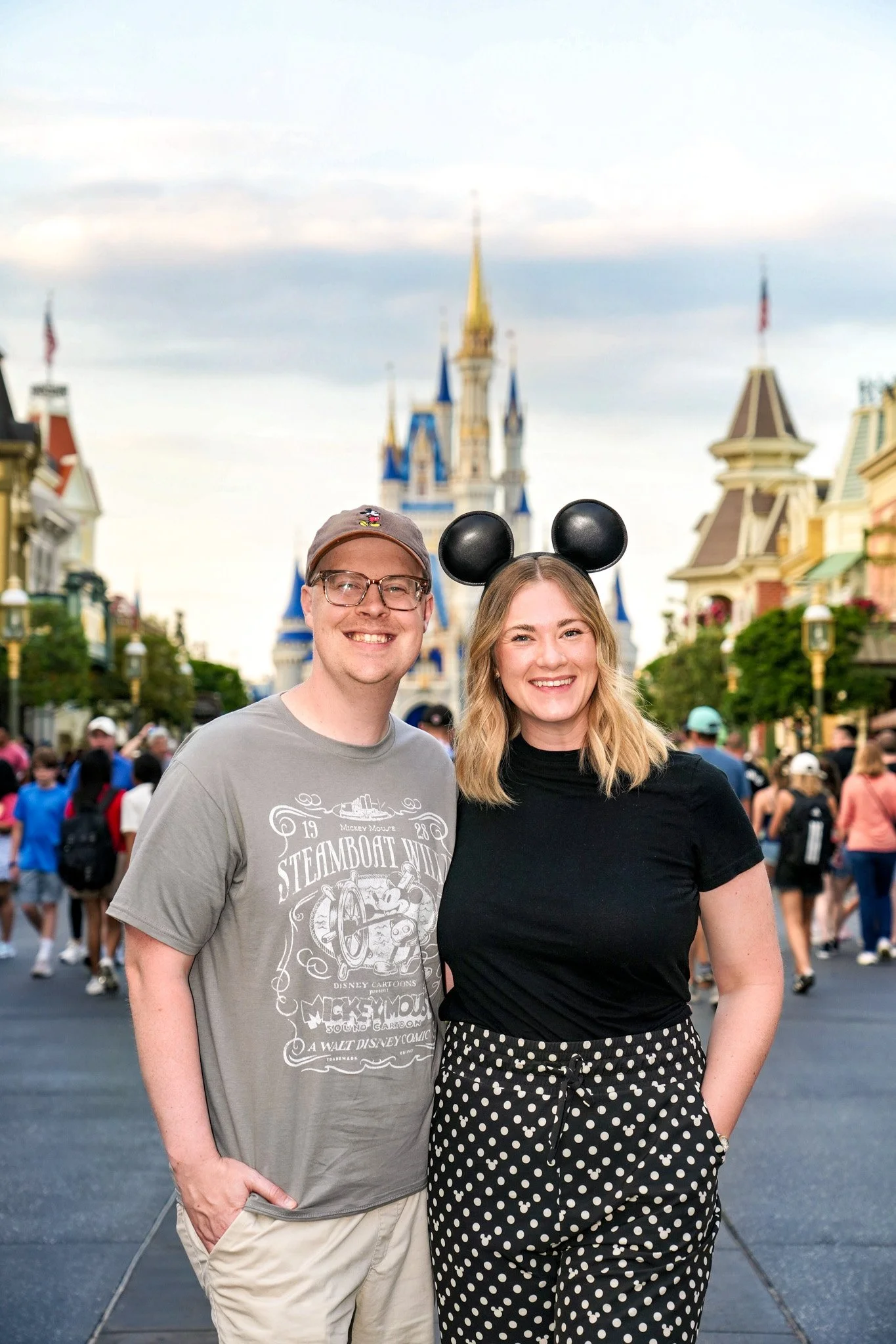 A smiling man and woman standing close together at a Disney theme park, with the castle in the background and a crowd of people around them.