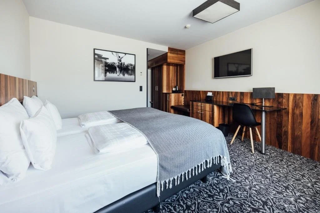 Modern hotel room featuring a neatly made bed with white linens and a gray throw, a wall-mounted TV, a wooden desk, and a black chair.