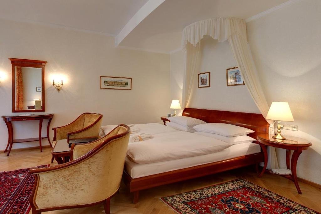 A hotel room with a large bed with a wooden headboard, cream curtains, two bedside lamps, vintage chairs, a mirror, framed pictures on the wall, and a patterned rug.
