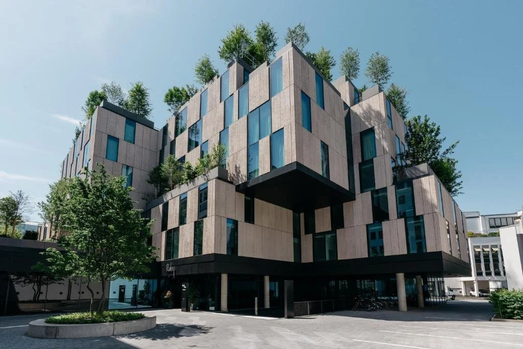 Modern multi-story building with wooden exterior panels and large glass windows, featuring greenery on upper levels and trees nearby.