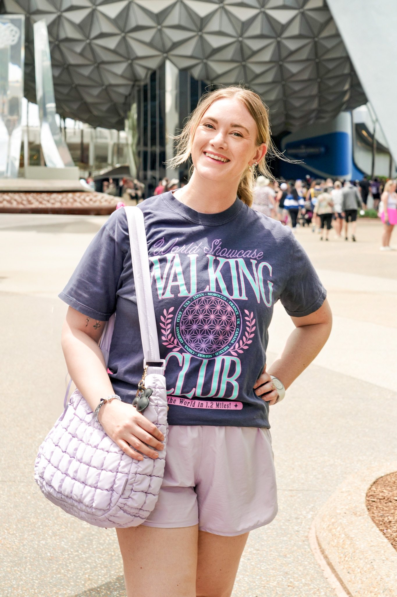 A young woman with light skin and blonde hair, smiling, standing outdoors in front of a modern building with a geometric facade and a crowd of people in the background. She is wearing a dark T-shirt with colorful text and design, light-colored shorts, and carrying a quilted pink bag and a white shoulder bag.