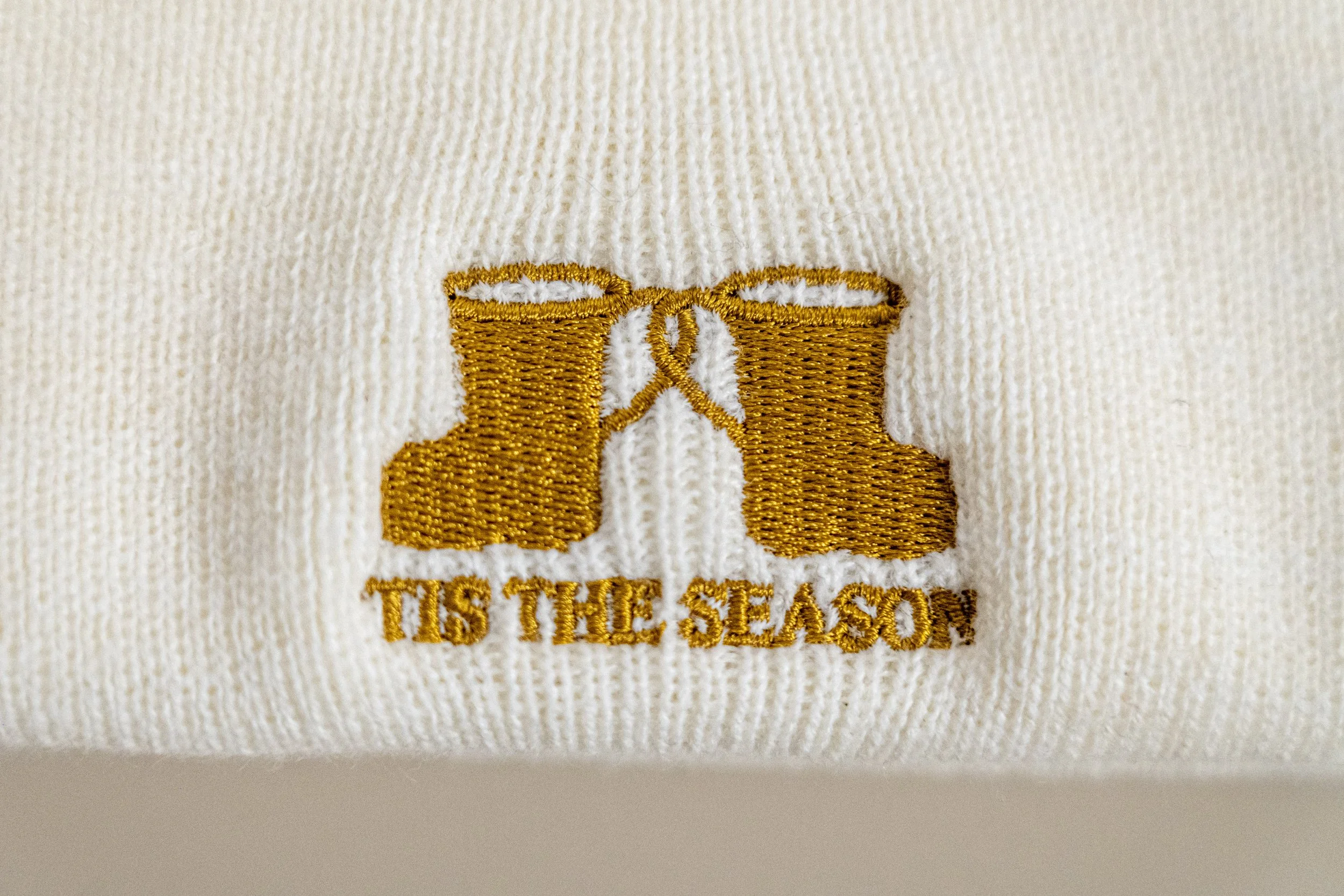 Close-up of cream-colored fabric with embroidered gold winter boots and the text "TIS THE SEASON" below.