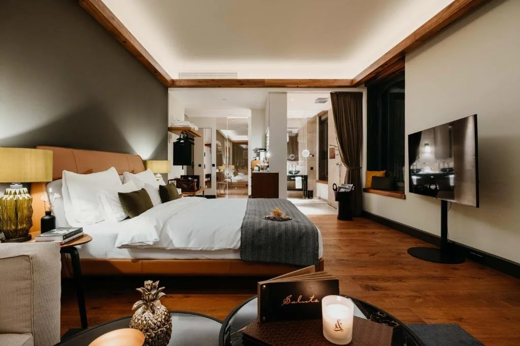 Well-lit hotel bedroom with a large bed, bedside lamps, TV, and a sitting area, with a view into the bathroom and additional seating in the background.