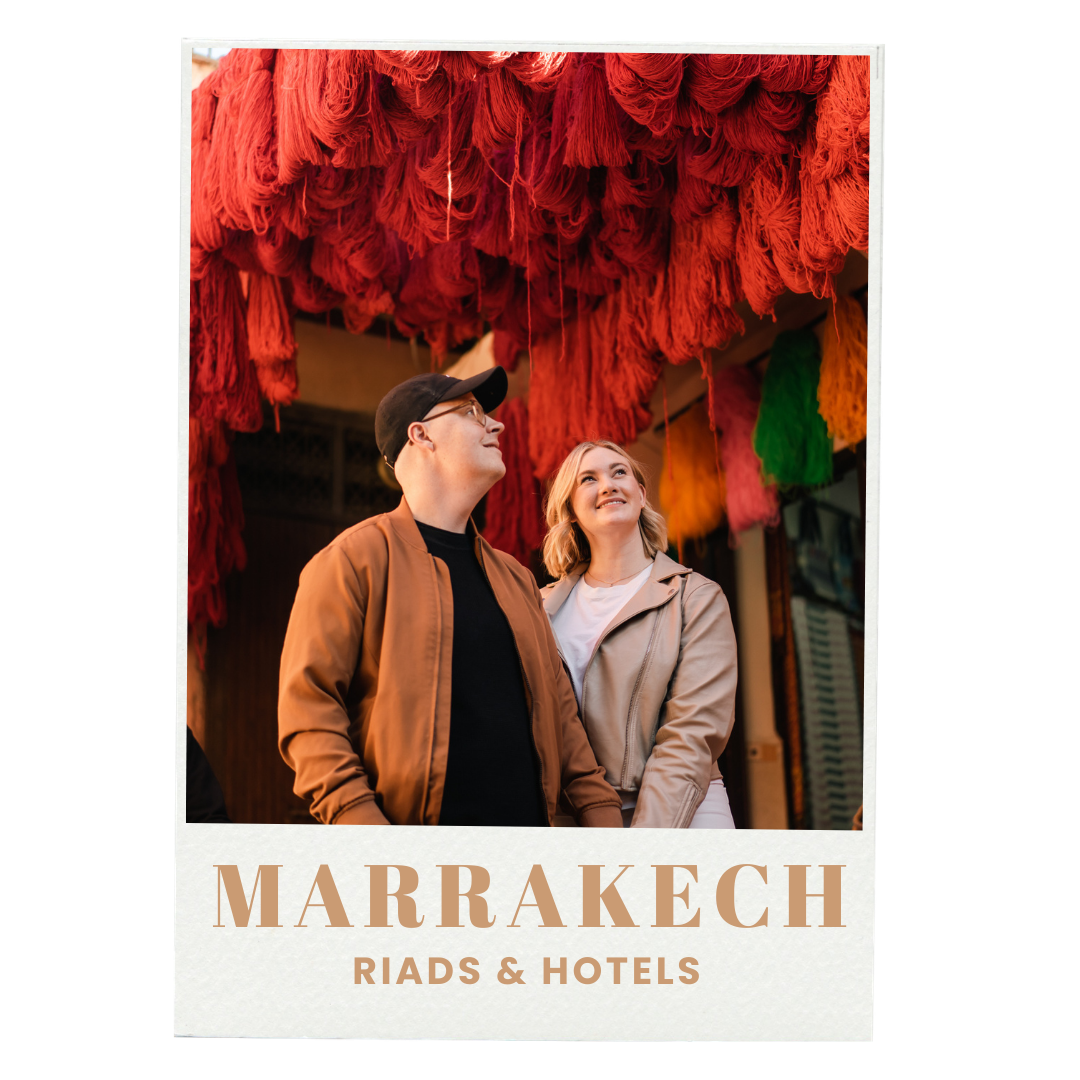 Two people, a man and a woman, standing underneath vibrant red and green hanging textiles during the daytime. The man is wearing glasses, a black cap, and a brown jacket, while the woman has blonde hair and is wearing a beige jacket. They are looking upwards with smiles. Text at the bottom reads "MARRAKECH RIADS & HOTELS."