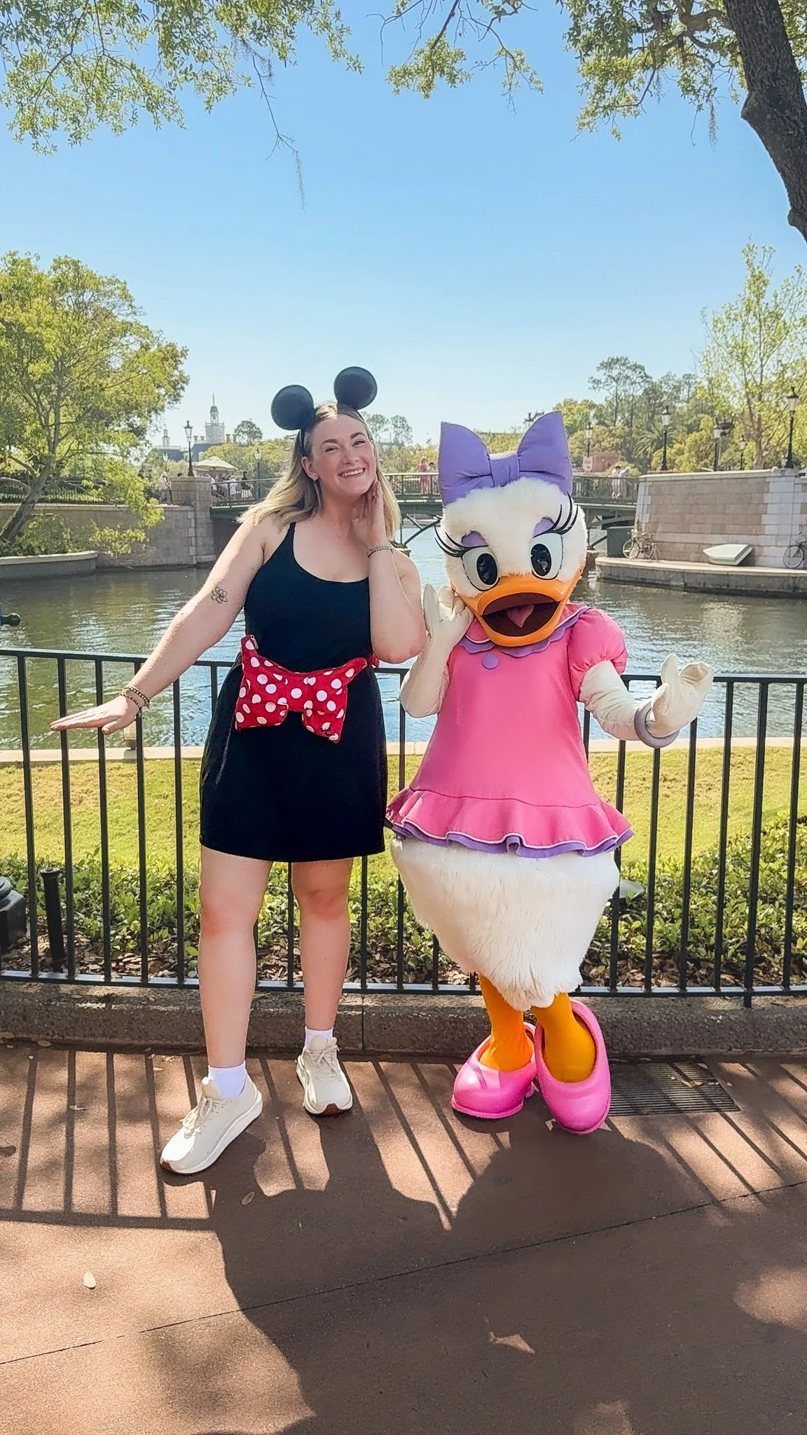 A woman wearing a black dress with a red polka dot bow and Mickey Mouse ears pose with a costumed Disney character resembling Daisy Duck dressed in a pink dress with a purple bow, standing by a waterfront on a sunny day.