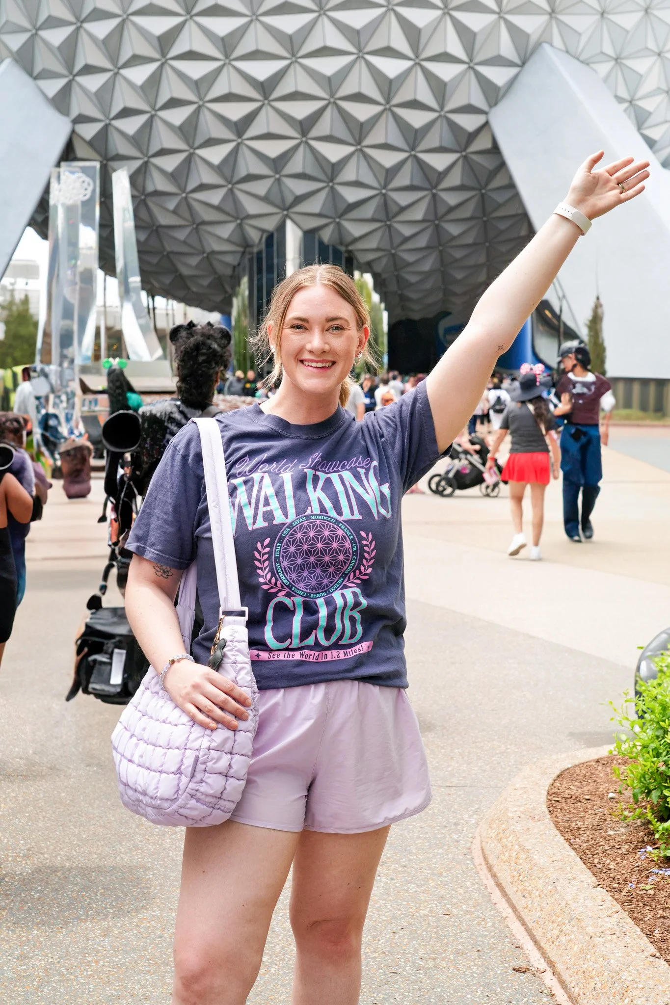 A young woman with blonde hair smiling at the camera, wearing a dark purple T-shirt and light pink shorts, standing outdoors in front of a large, modern building with a geometric dome. She has a quilted pink bag hanging from her left shoulder and is raising her right arm.